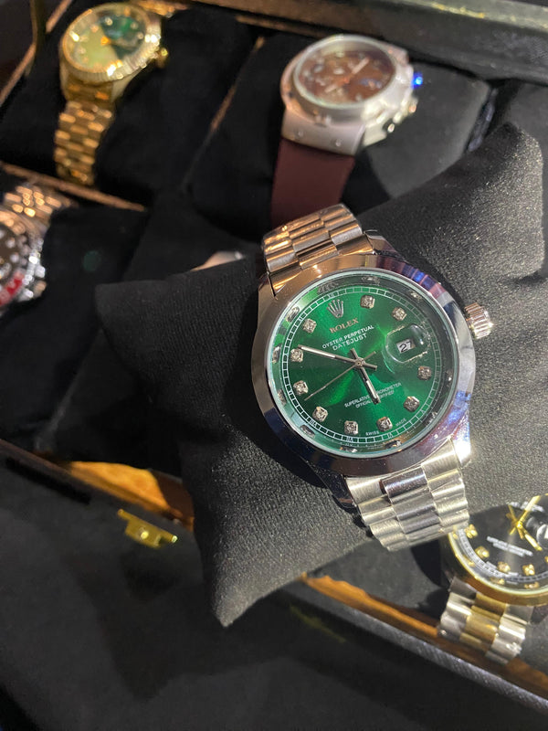 Luxury watch with green face and silver case on a black cushion, surrounded by other watches.